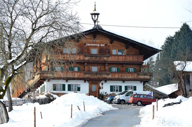 Ein traditionelles Holzhaus im Schnee mit mehreren Balkonen. Umgeben von einer winterlichen Landschaft und einigen Autos.