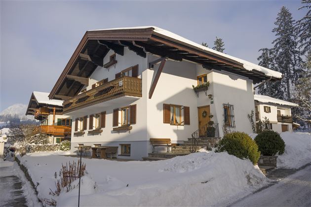 A traditional house in the snow with a steep roof and wooden balconies.  
Surrounded by a winter landscape and tall trees.