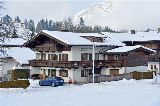 Ein gemütliches Haus im Schnee mit einem blauen Auto davor. Im Hintergrund sind schneebedeckte Berge und Nadelbäume zu sehen.