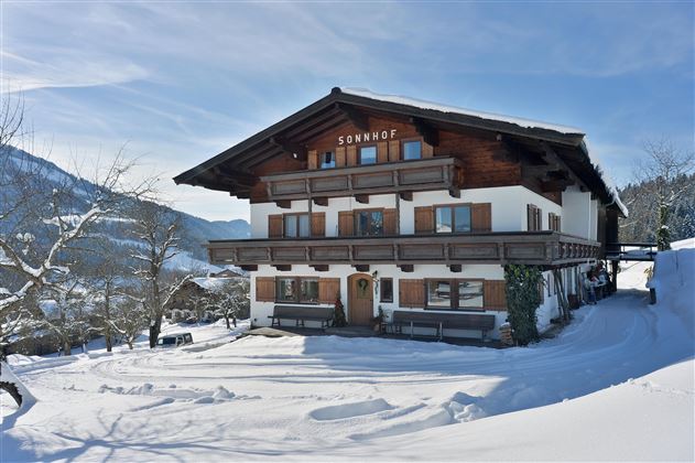 Ein gemütliches Holzhaus mit der Aufschrift "Bavaria" in einer verschneiten Landschaft. Im Hintergrund sind Berge und eine Kirche zu sehen.