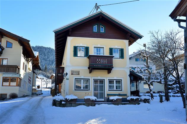Ein charmantes, zweigeschossiges Haus im Schnee, umgeben von verschneiten Bäumen. Die Fassade ist traditionell mit Fenstern und einem Balkon gestaltet.