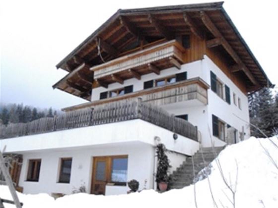 Ein modernes Haus mit Holzdetails, umgeben von schneebedecktem Boden und Bergen im Hintergrund. Der Himmel ist klar und die Umgebung wirkt ruhig.