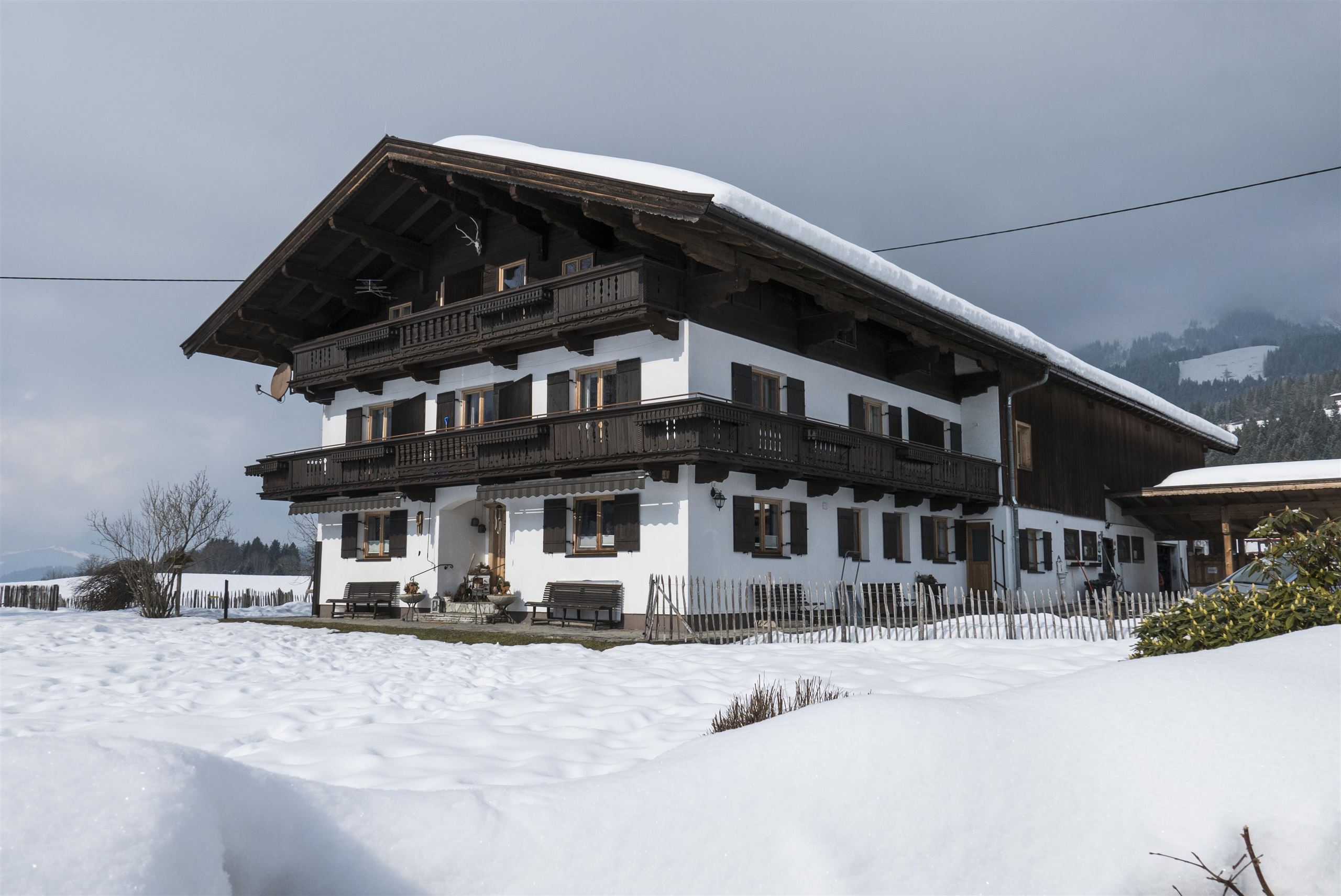 Een traditioneel alpenhouthuis in de winter, omgeven door sneeuw. Op de achtergrond zijn sneeuwbedekte bergen zichtbaar.
