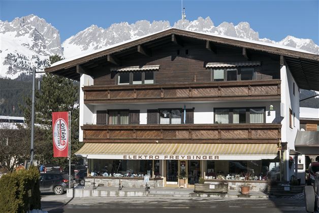 Ein traditionelles Holzgebäude mit einem Geschäft namens Elektro Freydinger. Im Hintergrund sind schneebedeckte Berge und ein klarer blauer Himmel zu sehen.