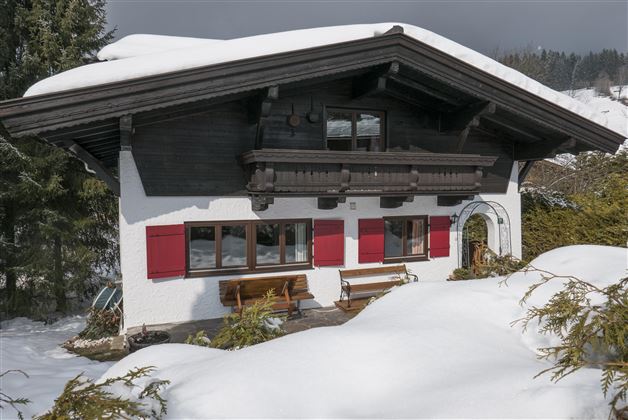 A charming alpine house with red shutters. Surrounded by snow-covered ground and mountains in the background.