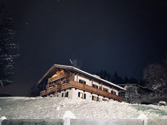 Ein gemütliches Haus im Schnee bei Nacht. Es umgeben Bäume und die Landschaft wirkt friedlich und winterlich.