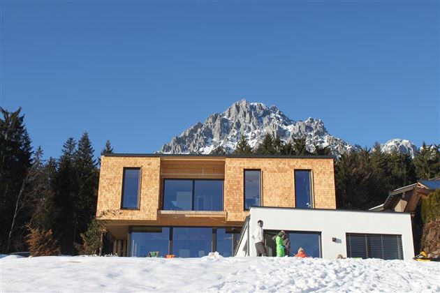 A modern house made of wood and concrete, surrounded by snow-covered mountains. The clear blue sky gives the scene a calm atmosphere.