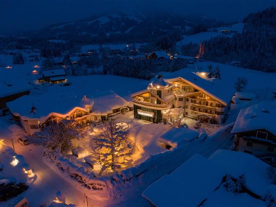 Ein malerisches Hotel in einer verschneiten Landschaft bei Nacht. Das Gebäude ist warm erleuchtet und von schneebedeckten Bäumen umgeben.