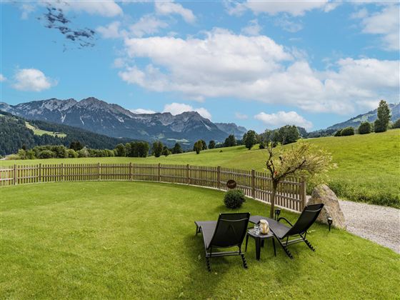 A beautiful meadow with two loungers and a view of the mountains. The sky is bright blue with some clouds.