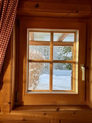 A window with a wooden frame that overlooks a snowy landscape. The gentle sun rays bathe the room in warm light.