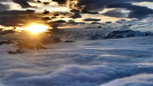 A sunrise over snow-covered mountains. The sky is filled with dramatic clouds and a sea of mist.