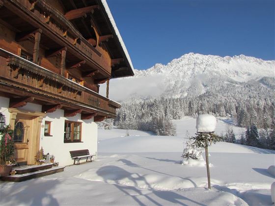 Ein gemütliches Chalet im Winter, umgeben von viel Schnee. Im Hintergrund erheben sich majestätische Berge unter einem klaren Himmel.