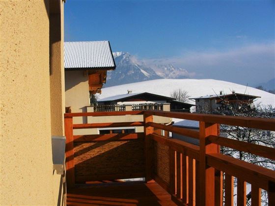 A balcony with a wooden railing that offers a picturesque view of snow-covered mountains. The sky is clear and blue, which impressively highlights the winter landscape.