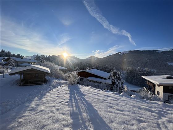 A snowy landscape with small wooden houses. The sun rises behind the mountains and the sky is clear.