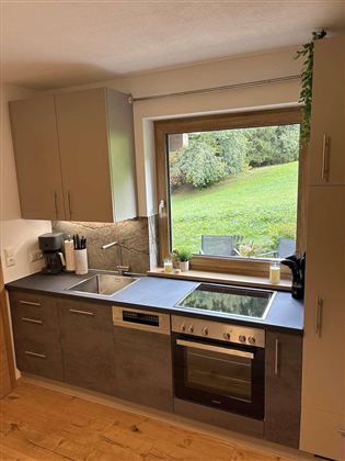 Eine moderne Küche mit einer großen Fensterfront, die einen Blick auf den Garten bietet. Der Raum ist hell und gut ausgestattet mit Kochgeräten.