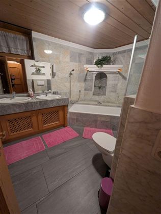 Ein modernes Badezimmer mit einer Badewanne und Dusche. Die Wände sind mit Steinfliesen verkleidet, und pinkfarbene Teppiche sorgen für einen farbenfrohen Akzent.