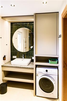 A modern bathroom with a round mirror and a sink. On the right, there is a washing machine in a simple cabinet.