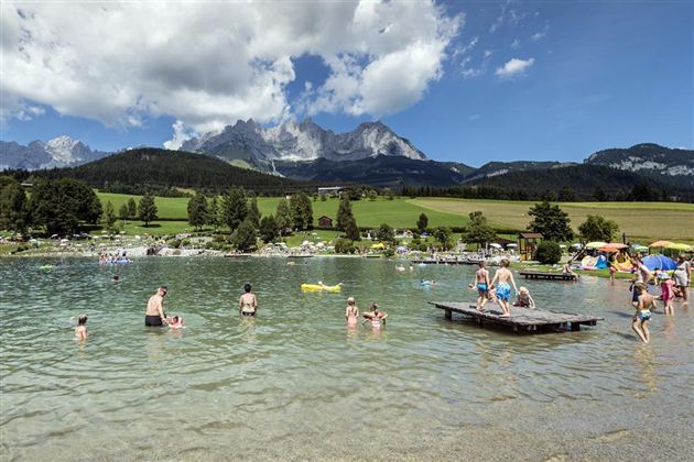 A clear lake surrounded by green meadows and mountains in the background. People are enjoying the water and the sun.