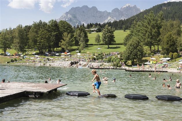 A boy is jumping on foam islands in the water. In the background, swimmers and a green meadow with mountains can be seen.