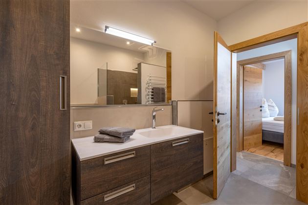 Ein modernes Badezimmer mit Holzschränken und einem hellen Spiegel. Im Hintergrund ist ein weiterer Raum zu sehen.