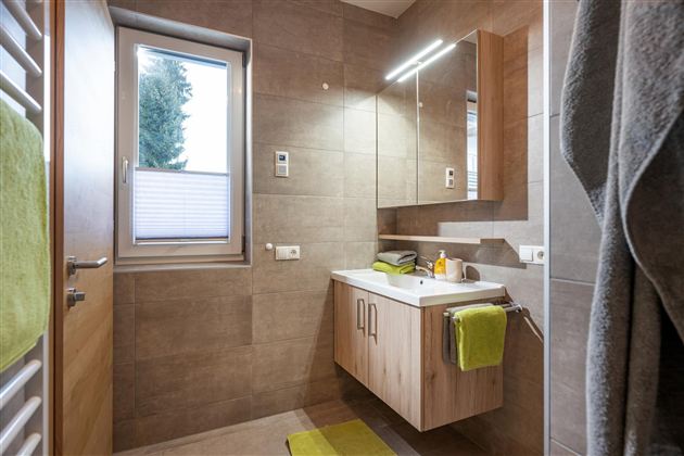 A modern bathroom with a wood vanity and a large mirror. Light gray tiles and green towels create a fresh atmosphere.