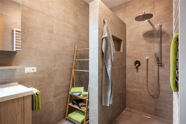 Modern shower with gray tiles and a rectangular shower area. A towel rack and toiletries are visible in the room.