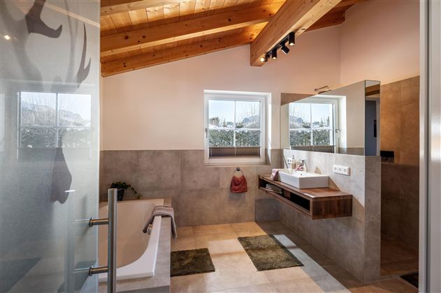 Ein modernes Badezimmer mit Holzdecke und Fenster, das viel Tageslicht bietet. Die elegante Ausstattung umfasst ein Waschbecken, einen großen Spiegel und eine Badewanne und grosszügige begehbare Dusche.