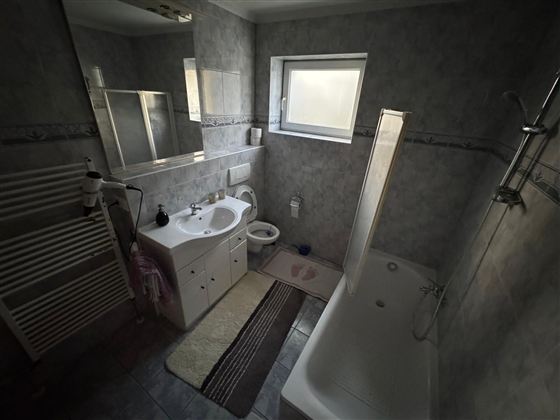 Ein modernes Badezimmer mit einer Badewanne, einer Dusche und einem Waschbecken. Die Wände sind in hellen Farben gehalten und es gibt ein Fenster für Tageslicht.