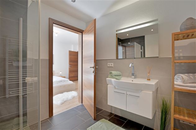 Ein modernes Badezimmer mit Dusche, Waschbecken und großer Spiegelleuchte. Die Tür öffnet sich zu einem angrenzenden Schlafzimmer.