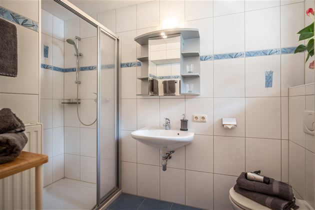A modern bathroom with a shower, sink, and a mirror. The walls are tiled, and there are towels in dark colors.