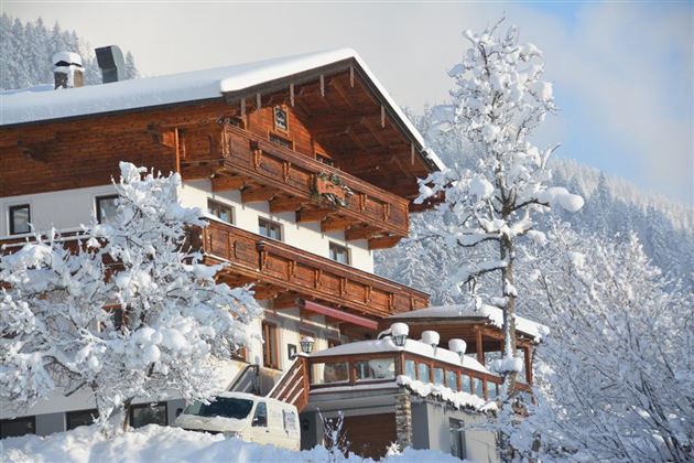 Ein schneebedecktes Gebäude im alpinen Stil umgeben von winterlichen Bäumen. Der Himmel ist klar und die Berge sind im Hintergrund sichtbar.