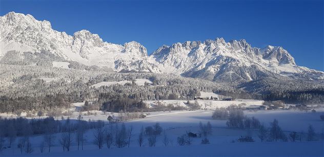 An impressive winter landscape with snow-covered mountains and a clear blue sky. In the foreground, a quiet, snowy plain stretches out.