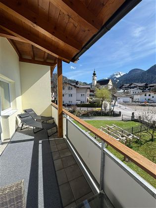 A balcony with loungers and a view of a picturesque village in the mountains. The surroundings are green and the sky is clear.
