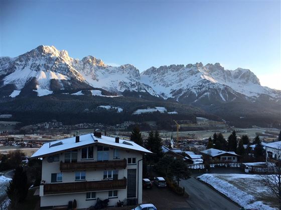 A picturesque mountain panorama with snow-capped peaks and a clear sky. In the foreground, a few houses and trees can be seen.