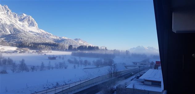 A winter landscape with snow-covered mountains and a clear blue sky. In the foreground, there is a road and some buildings visible.