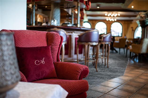 Ein gemütlicher Sessel mit einem Kissen in rot steht in einem stilvollen Restaurant. Im Hintergrund sind elegante Stühle und eine Bar sichtbar.
