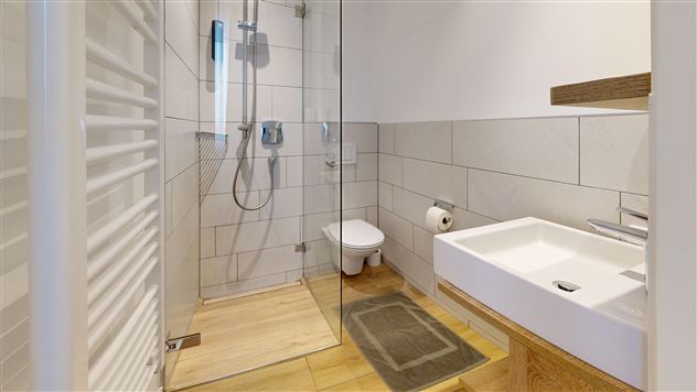 A modern bathroom with a glass shower and a white sink. The floor is made of wood and the walls are tiled in light colors.