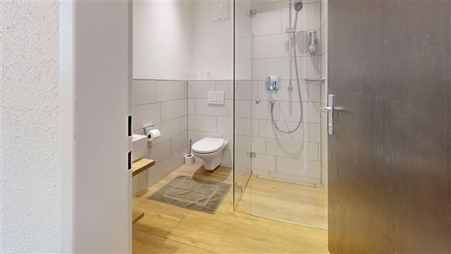 A modern bathroom with shower and toilet. The floor is made of wood and the walls are covered in light tiles.