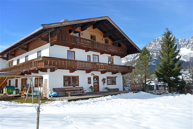 Ein traditionelles Haus im alpine Stil, umgeben von Schnee. Im Hintergrund sind Berge und ein blauer Himmel zu sehen.