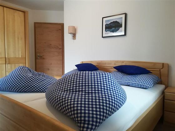 A cozy bedroom with a double bed and large checked cushions. The walls are light and there is a wooden wardrobe as well as a picture on the wall.