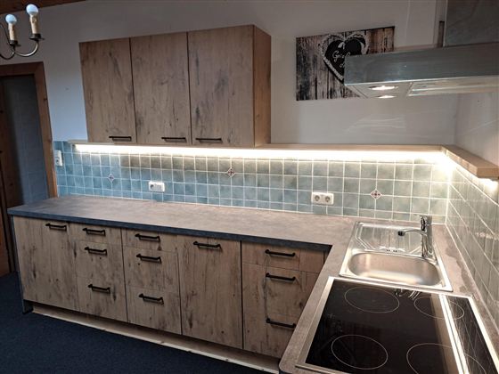 Eine moderne Küche mit Holzschränken und einer grauen Arbeitsplatte. Die Wände sind mit blauen Fliesen ausgestattet und es gibt eine Kochplatte sowie ein Spülbecken.