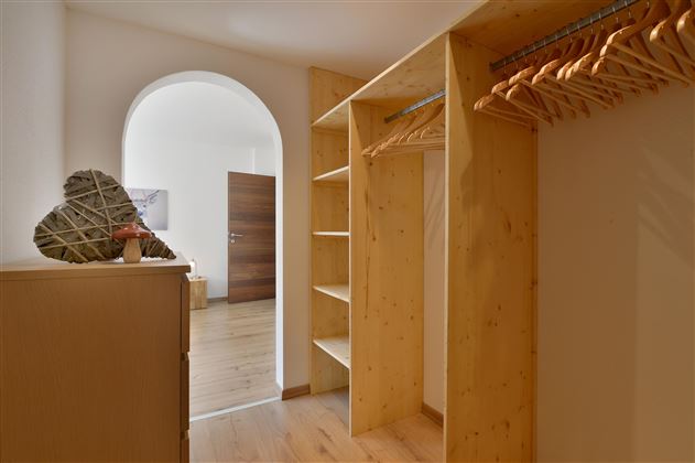 Ein heller Eingangsbereich mit einem offenen Kleiderschrank aus Holz. Im Hintergrund ist eine Tür sichtbar, die zu einem weiteren Raum führt.