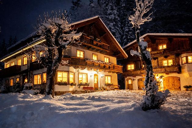 A cozy, illuminated chalet in the snow at night. The surroundings are wintry and idyllic with snow-covered trees.