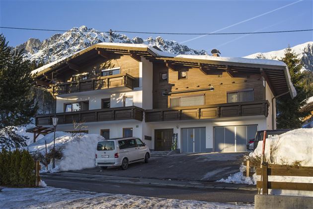 A modern wooden house in a snow-covered landscape with mountains in the background. There is a parking lot and clear blue skies.