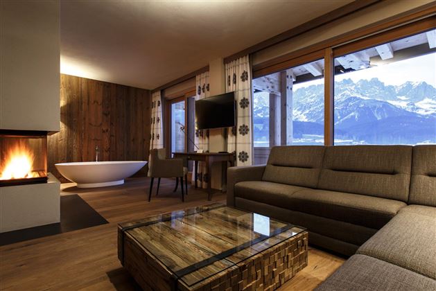 A modern living room with a cozy sofa, a fireplace, and a table.  
Large windows provide a view of the mountains and let light in.