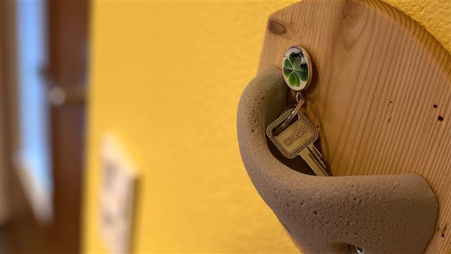 Een sleutel hangt aan een houten houder aan een gele muur. De scène toont een eenvoudige en uitnodigende inrichting.