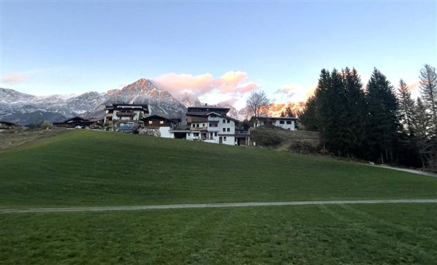 Een pittoresk landschap met huizen op de voorgrond en bergen op de achtergrond. De lucht is helder en de wolken kleuren rood.