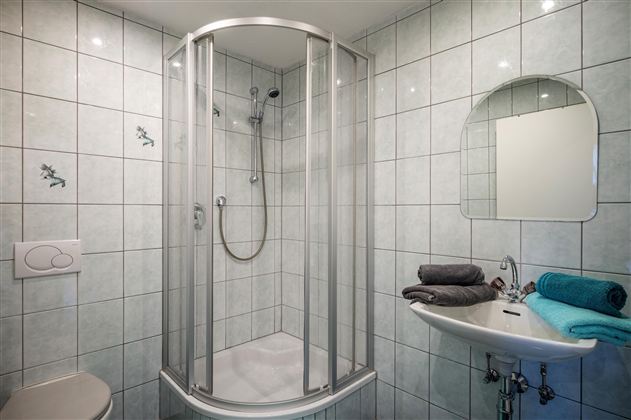 Ein modernes Badezimmer mit einer Dusche aus Glas und weißen Fliesen. Handtücher in Blau und Grau liegen auf dem Waschbecken.