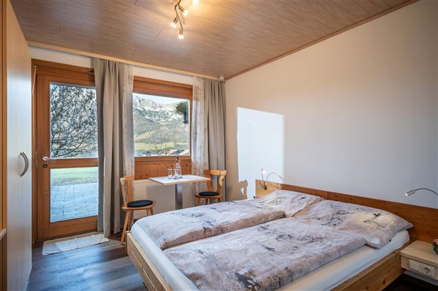 Ein gemütliches Schlafzimmer mit einem Doppelbett und Holz-Elementen. Bei Tageslicht ist ein Tisch mit zwei Stühlen sichtbar, und große Fenster bieten einen Blick nach außen.
