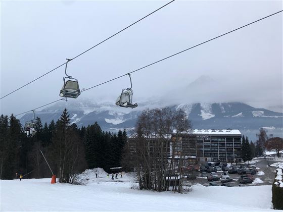 A snowy landscape with ski lifts in the background. Densely wooded mountains are visible in the distance.